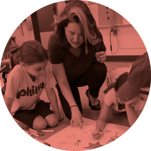 An image of a woman sitting with three young children and helping them as they color and create a picture with their crayons.