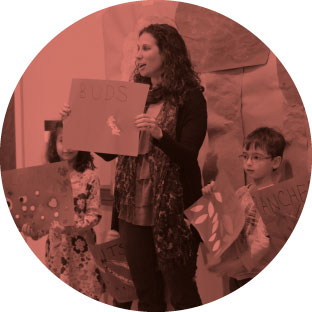 A woman who looks to be passionate and enthusiastic standing next two young children as they all hold up pictures and posters that they each created. They are showing them to an audience and speaking about them.