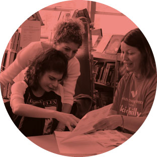 An image of two women, smiling and enthusiastically helping a young girl complete a worksheet. They are in a library or setting that has a lot of books behind them. One of the women is sitting next to her and the other is behind her with her arm around the girl