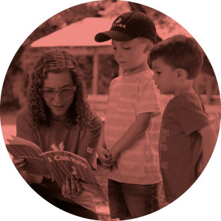 A woman with curly hair and glasses smiling as she kneels next to two young boys, one wearing a baseball cap and reads a book to them.