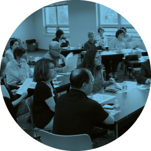 A picture of nine adults sitting at tables with papers in front of them, all facing the same way, learning. They all look intrigued and focused to continue listening to what they are learning about.