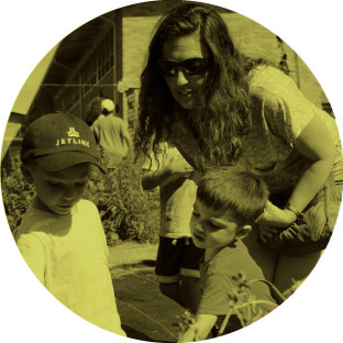 A woman with sunglasses bends over to talk to two young children who are playing, one of them wearing a baseball cap.