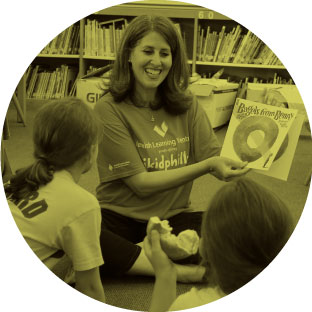 A woman wearing a jkidphilly shirt, smiling as she enthusiastically reads a book to young children sitting in front of her.