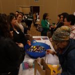 A group of people standing around a table with food and supplies, smiling and making conversation with each other.