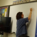 A woman writing on a whiteboard, teaching others. She has short, curly hair and is wearing a short sleeve blue shirt.