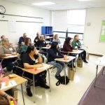 A group of adults, both men and women, sitting at desks in a classroom with papers in front of them, learning.