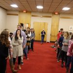 A picture of a group of people standing in lines, ready to learn or play a game. There are wood doors in the back and a red carpet.