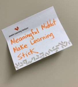 A sticky note with the words “Meaningful Middot Make Learning Stick” written in orange letters on it. At the bottom of the sticky note are grey Hebrew letters.