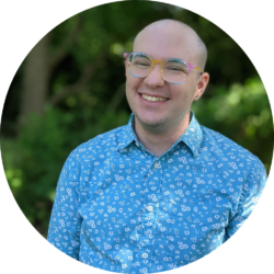 A white non-binary person with a shaved head and in a turquoise shirt, rainbow glasses, and heart earrings stands smiling in front of a blurred green background.