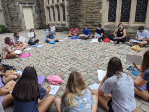 A photograph of about 30 teens wearing masks sitting in a circle on the ground next to the side of a building made out of stone. Some of the teens are holding up whiteboards with writing on them. 