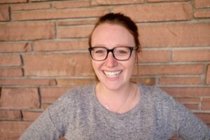 A headshot of Carly Coons, a smiling redhead with large black-rimmed glasses in front of a brick wall.