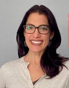 A headshot of Aimee Kramer, a brunette with long, wavy hair and dark red lipstick, in front of a grey background.