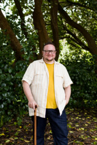 A photo of Rabbi Elliot Kukla, who is standing casually with a big smile in the middle of a grove of trees. He is using a cane.